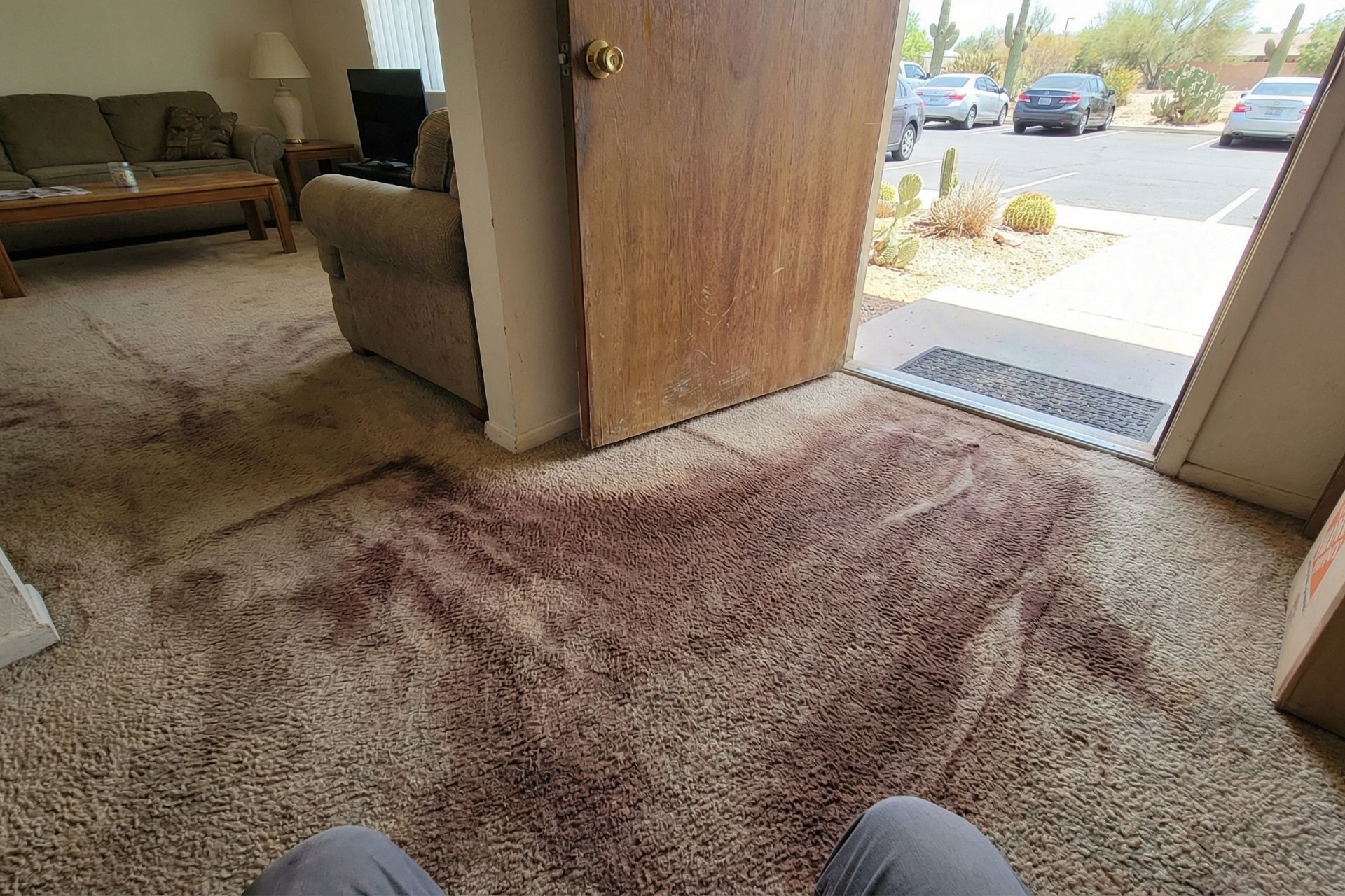 First-person view of heavily soiled beige carpet in rental property entryway showing large dark brown dirt stains and traffic patterns near open front door, with furnished living room visible in background and desert landscaping with parked cars outside.