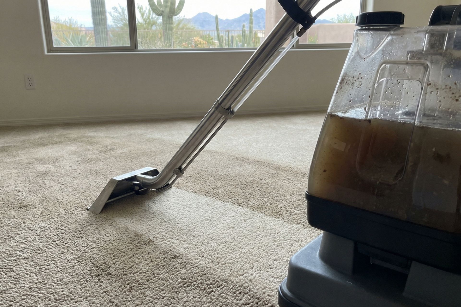 Professional carpet cleaning equipment in action showing a steam cleaning wand with metal extension tube on beige carpet, with a recovery tank containing dirty brown water visible in the foreground, and desert mountain landscape with saguaro cacti visible through windows.